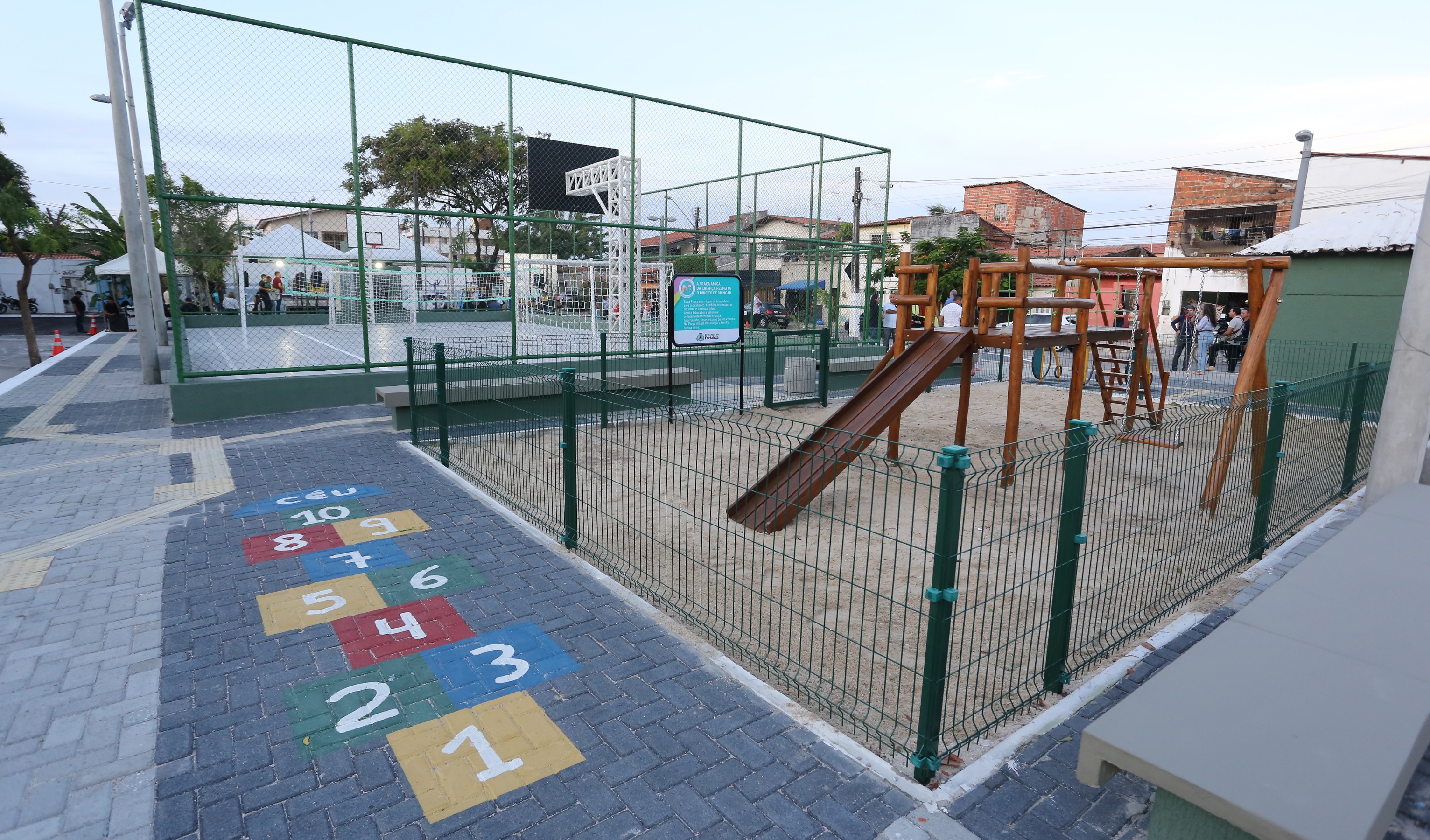 praça reformada com amarelinha pintada na calçada, parquinho infantil e quadra esportiva